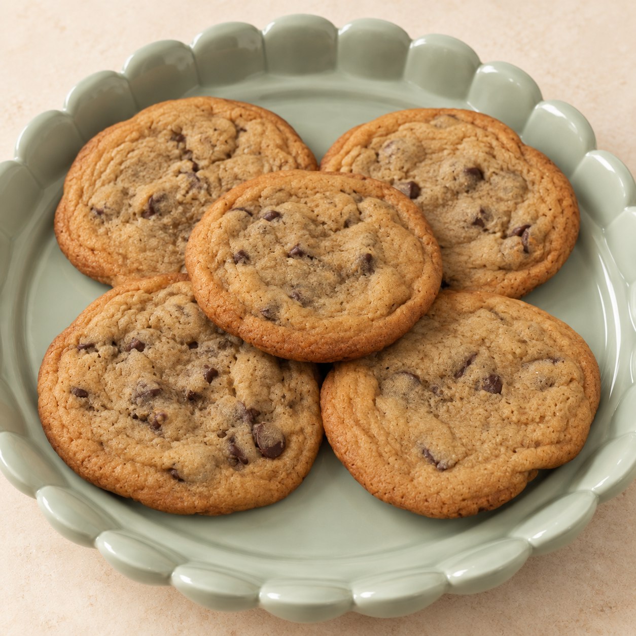 Five golden-brown chocolate chip cookies overlapping on a pale green scalloped platter.